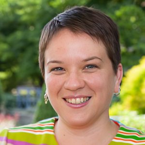 Person smiling outdoors with greenery in the background, wearing hoop earrings and a colorful striped shirt.
