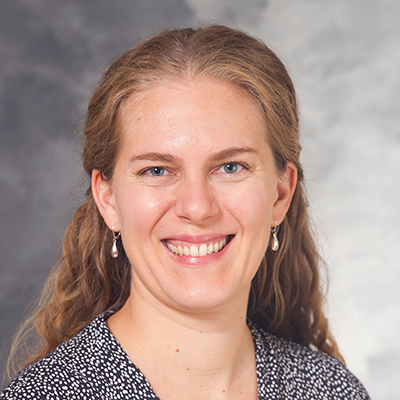 Headshot of a smiling person with long blonde hair, wearing a dark patterned blouse and small dangling earrings.
