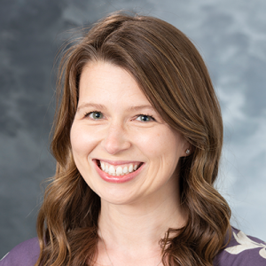 Portrait of a smiling person with long, wavy brown hair against a gray, cloudy background.