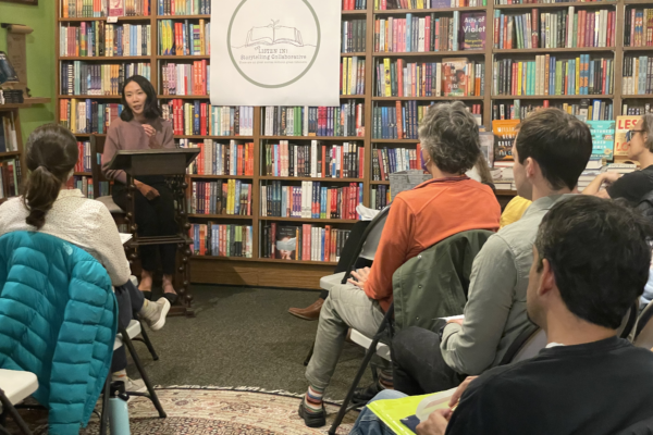Woman speaking at a podium in a bookstore with an audience seated in folding chairs.