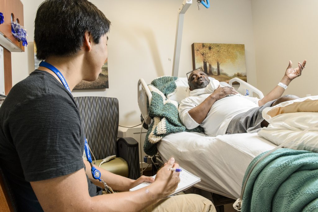 A medical professional takes notes while conversing with a patient lying in a hospital bed.
