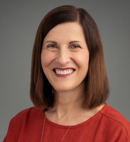 Headshot of a person with shoulder-length brown hair, wearing a red top, and smiling against a gray background.