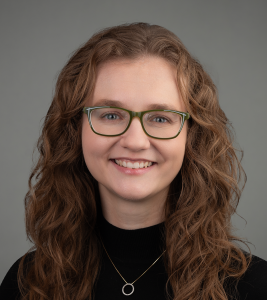 Headshot of a person with wavy brown hair, green glasses, and a black top, smiling in front of a grey background.