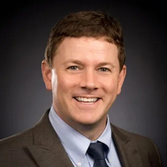 Headshot of a man with short brown hair, smiling, wearing a dark gray blazer, light blue dress shirt, and navy tie, against a dark gray background.