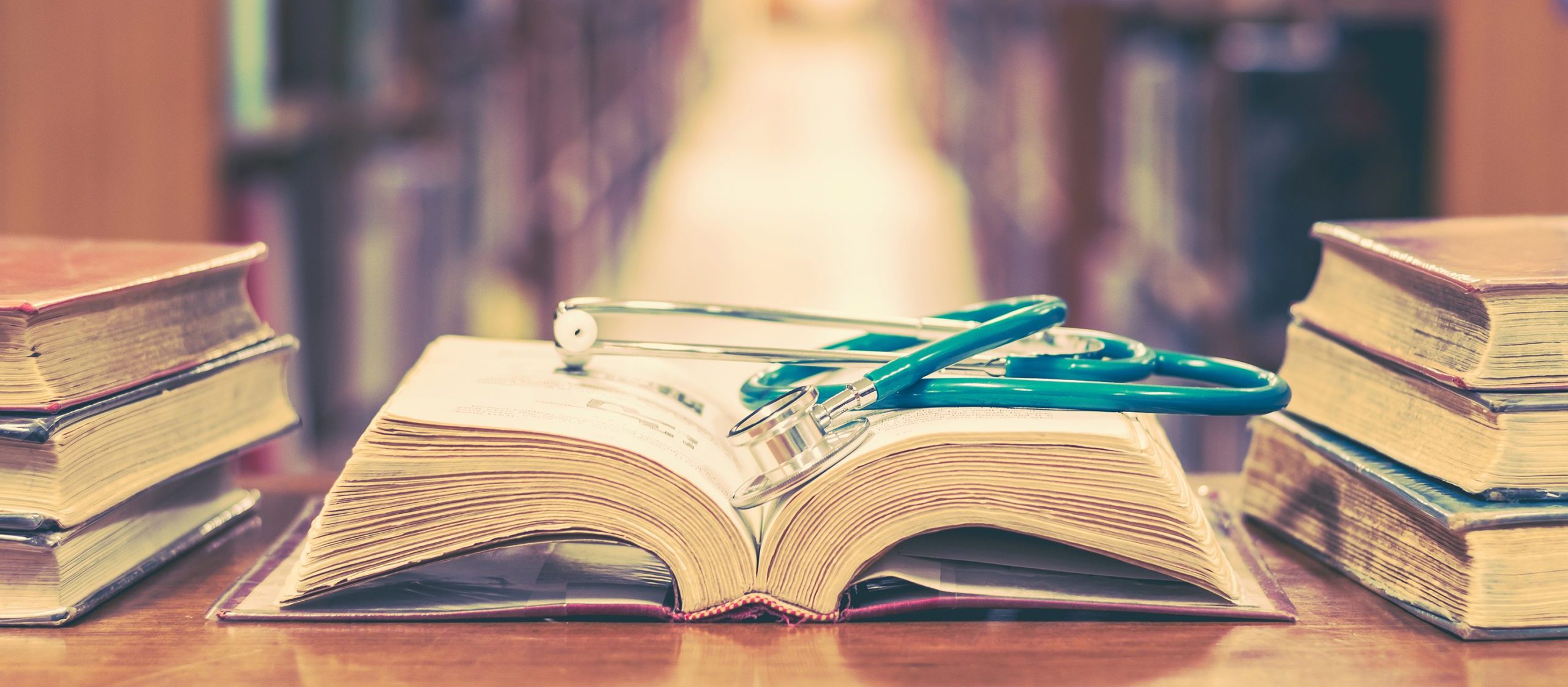 Old book in library with stethoscope on open textbook, stack piles of texts on reading desk, and aisle of bookshelves in study class room background for medical school education learning concept