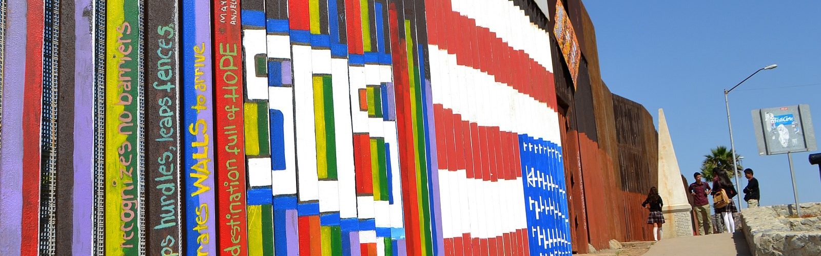 Colorful murals with messages of hope on the U.S.–Mexico border wall, people gathered nearby under blue sky.