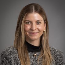 Headshot of a woman with long straight brown hair, smiling, wearing a gray tweed blazer over a black turtleneck, against a neutral gray background.