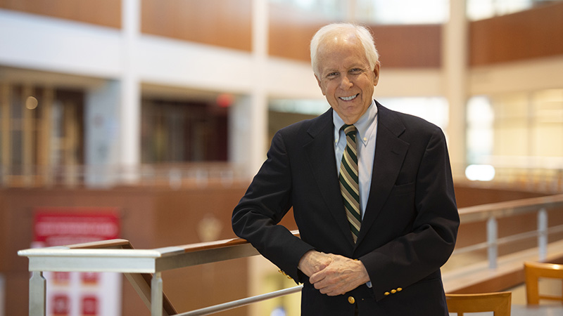 
An older man with white hair, wearing a dark navy blazer, a light blue button-down shirt, and a gold and navy striped tie. He is smiling and leaning against a railing in a bright, multi-level indoor atrium with warm wood accents and a blurred background.