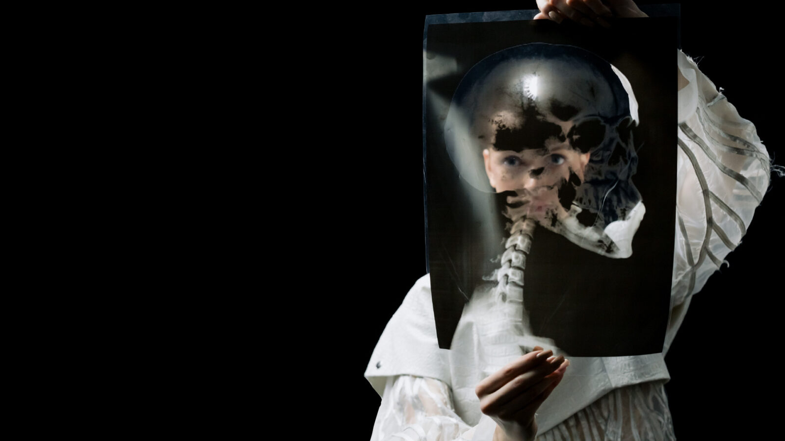 An person in textured white clothing holds a large X-ray transparency in front of their face against a solid black background. The X-ray, which shows a human skull and cervical spine, is positioned so the skeletal neck aligns with the person's body, while their real eyes and face are partially visible through the dark film.