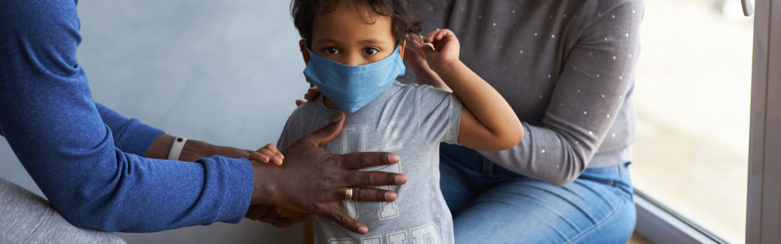 In this image, a young child wearing a blue cloth face mask is being helped by two adults, likely parents. One adult, wearing a blue long-sleeved shirt, has a hand on the child's chest, while the other, in a grey top and blue jeans, is visible in the background. The child is looking directly at the camera, and the scene appears to be set indoors near a window.