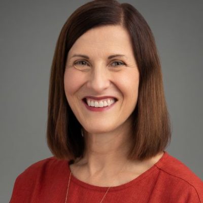 Headshot of a person with shoulder-length brown hair, wearing a red top, and smiling against a gray background.