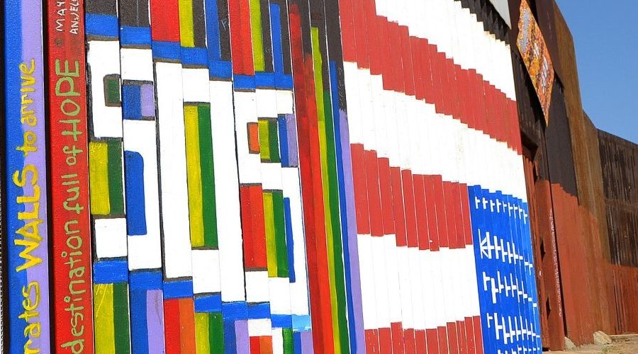 Colorful murals with messages of hope on the U.S.–Mexico border wall, people gathered nearby under blue sky.